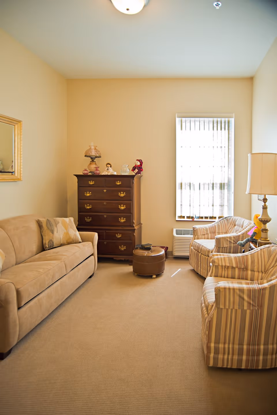 A cozy living room with a beige sofa on the left, two striped armchairs on the right, and a wooden chest of drawers against the back wall. The room has a window with vertical blinds, a small round ottoman in the center, and a floor lamp beside one of the armchairs. The walls are painted light beige, and the carpet is a matching neutral tone.