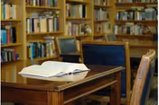 A wooden table with an open book on it, surrounded by chairs with blue cushions, in a room filled with bookshelves containing numerous books.