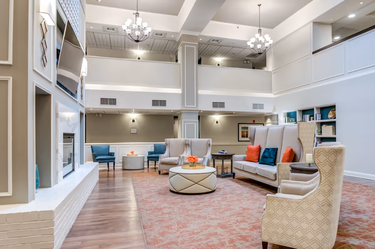 A spacious and well-lit living room area in a senior living facility featuring high ceilings with chandeliers, a large flat-screen TV mounted above a white brick fireplace, comfortable seating including armchairs and sofas with colorful cushions, a round ottoman coffee table with a flower arrangement, and built-in shelves with decorative items.