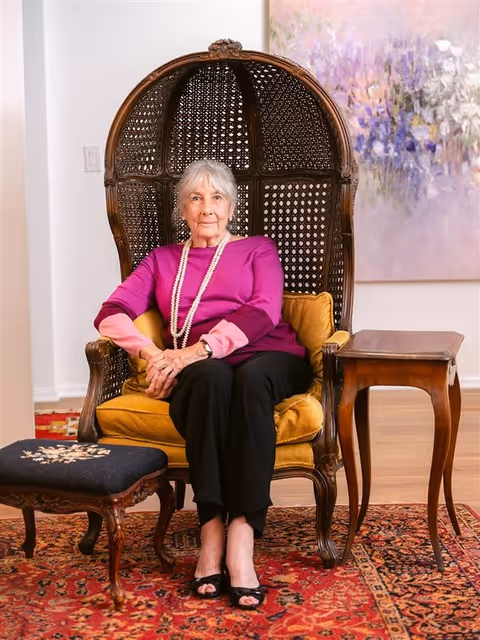 An elderly woman with gray hair wearing a magenta and pink sweater, black pants, and a long pearl necklace is seated in a large, ornate wicker chair with a yellow cushion. She is sitting in a room with a red patterned rug, a small wooden side table, and a matching footstool with a dark cushion. A large abstract painting with purple and white tones hangs on the wall behind her.