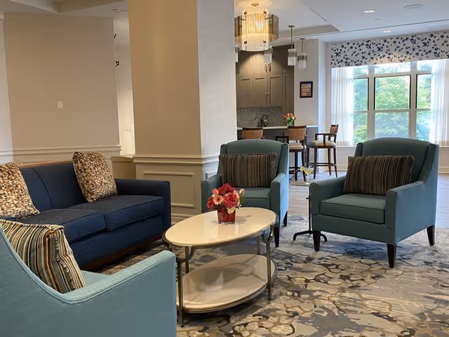 A cozy sitting area in a senior living facility with a dark blue sofa and two light blue armchairs arranged around a white oval coffee table with a small flower arrangement. In the background, there is a tall window with floral curtains, a high table with chairs, and cabinetry with pendant lights above.