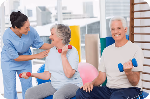 A female caregiver in blue scrubs assisting an elderly woman lifting red dumbbells while an elderly man sits nearby holding blue dumbbells in a bright exercise room with fitness equipment and large windows.
