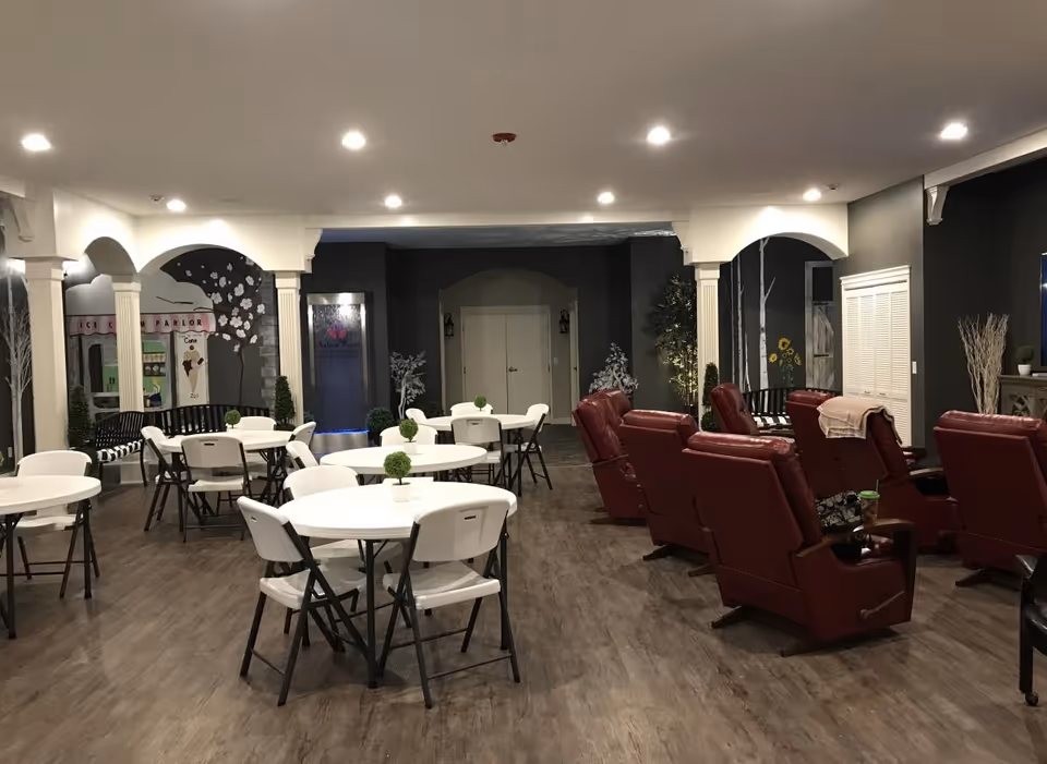 Interior view of a senior living facility common area with several round white tables and white folding chairs arranged on a wood floor. To the right, there are multiple red recliner chairs facing a TV or focal point outside the frame. The walls are decorated with murals, including a tree and an ice cream parlor scene. The ceiling has recessed lighting, and there are decorative columns and plants throughout the space.