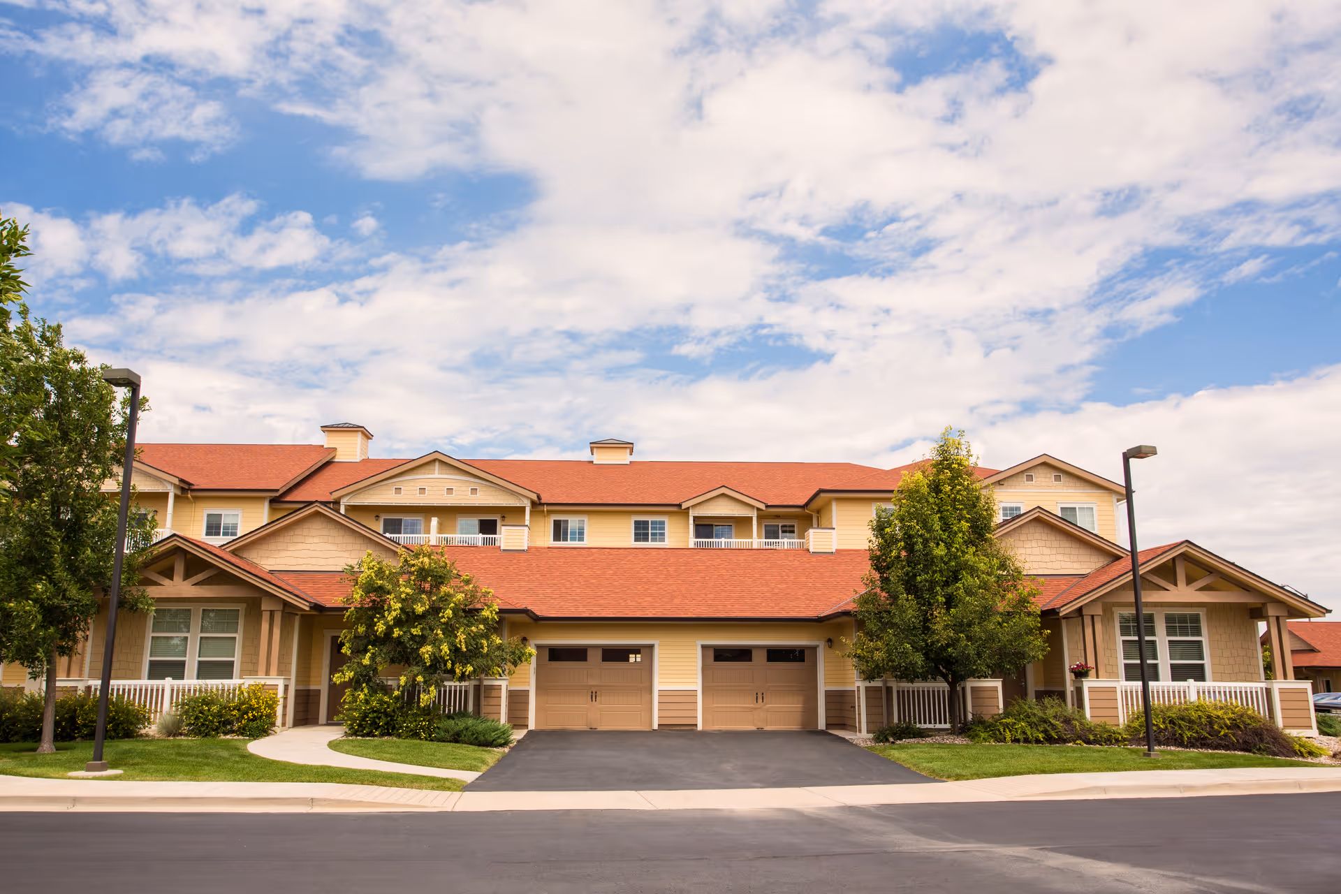 Front exterior view of a two-story senior living facility building with beige siding, red roof, two garage doors, trees, and a well-maintained lawn under a partly cloudy sky.