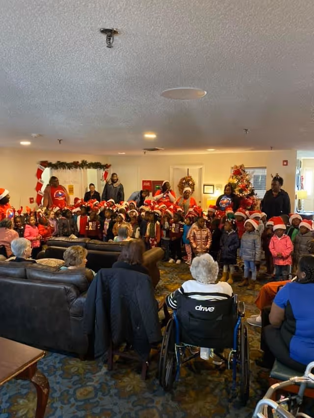 A group of children wearing Santa hats and festive clothing standing in a room decorated for Christmas, including a Christmas tree and garlands. Several elderly people are seated on couches and chairs, including one person in a wheelchair, watching the children. The setting appears to be a common area in a senior living facility.