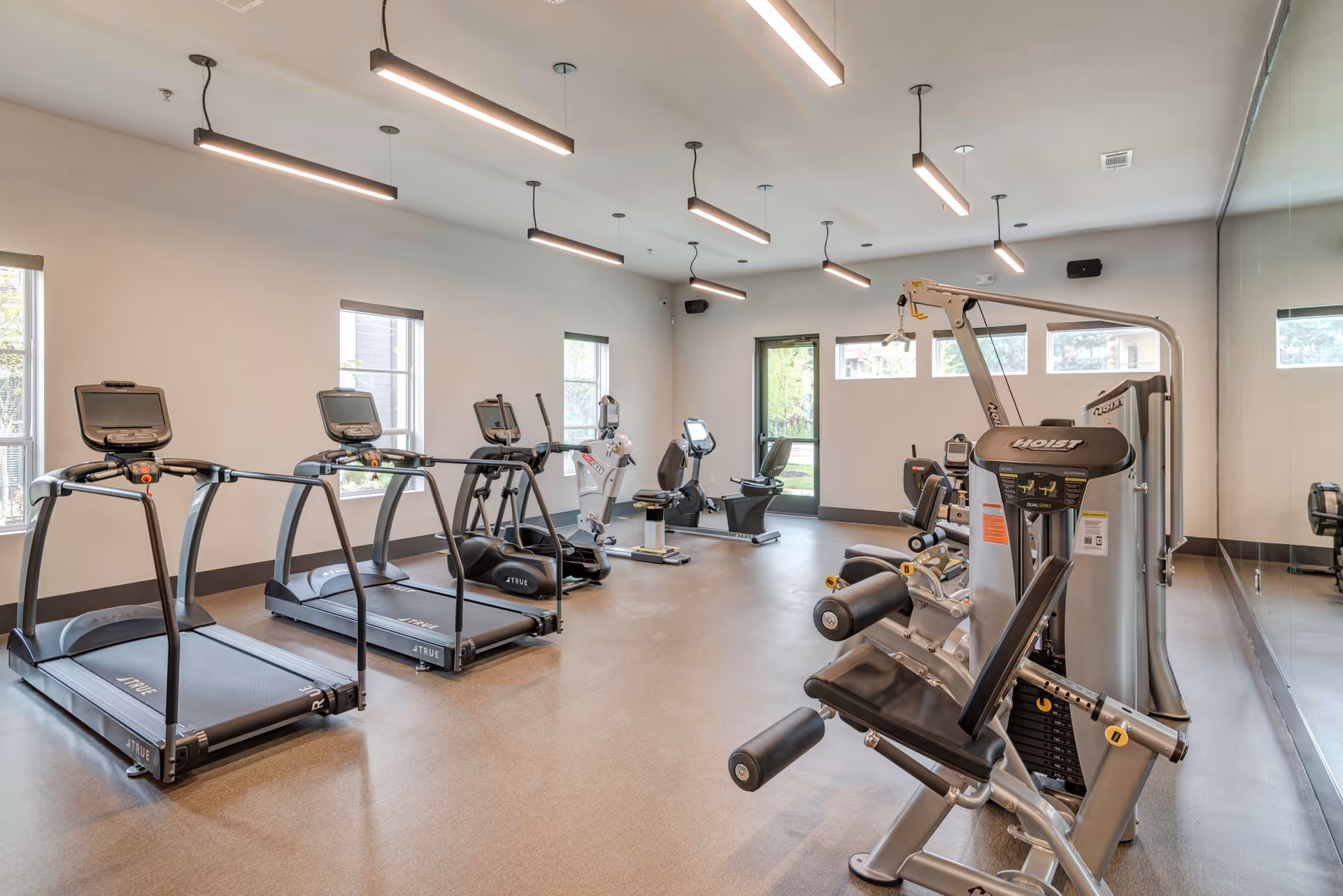 A bright and clean fitness room with several treadmills, exercise bikes, and weight machines. The room has large windows allowing natural light, modern ceiling lights, and a mirrored wall on the right side.