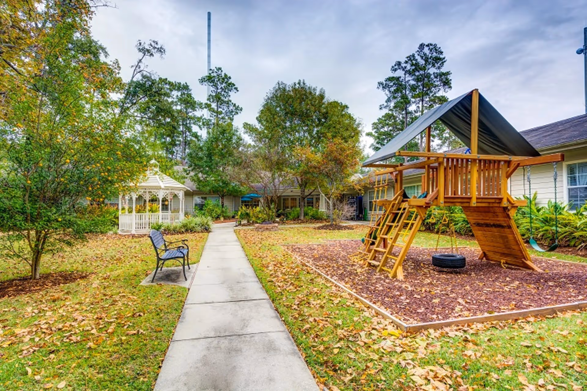Courtyard with a paved walkway leading past a bench to a white gazebo and a wooden playset, surrounded by trees and single-story buildings.