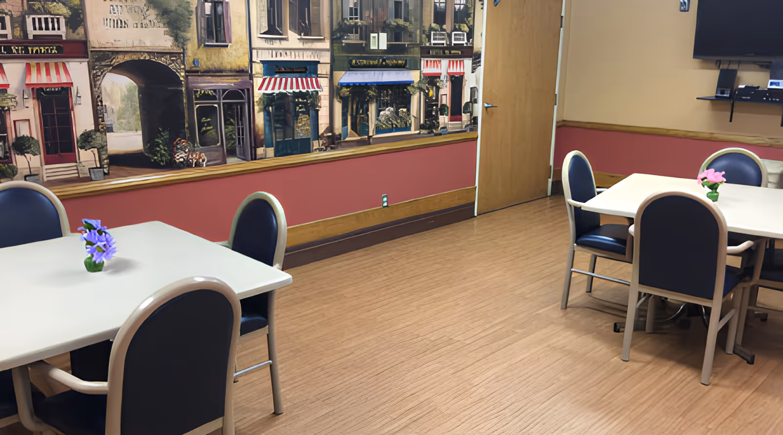Interior of a dining room with two tables, each surrounded by four chairs with dark blue cushions. Each table has a small vase with flowers, one with purple flowers and the other with pink flowers. The walls are painted beige and red with a mural depicting a street scene with buildings and storefronts. A wooden door and a mounted TV with shelves are visible on the right side.