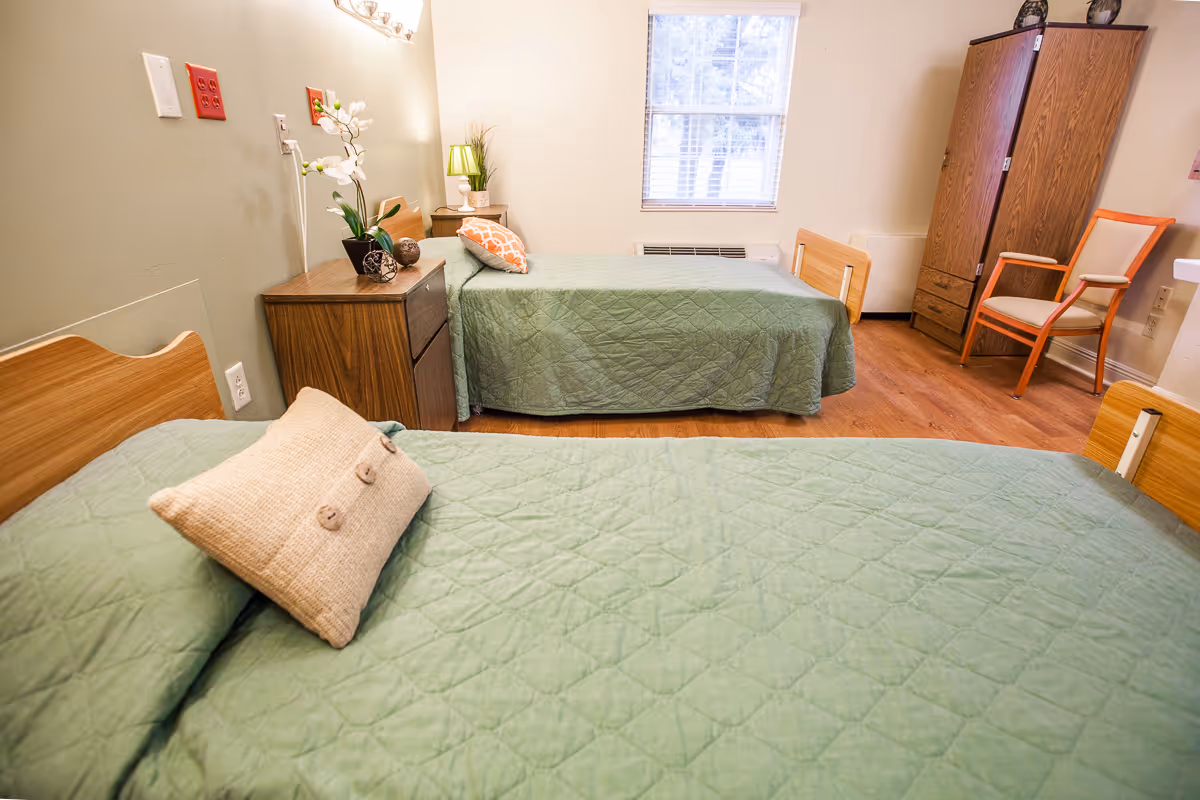 Two twin beds with green bedspreads in a furnished senior living bedroom with a nightstand, wardrobe, chair, and a window.