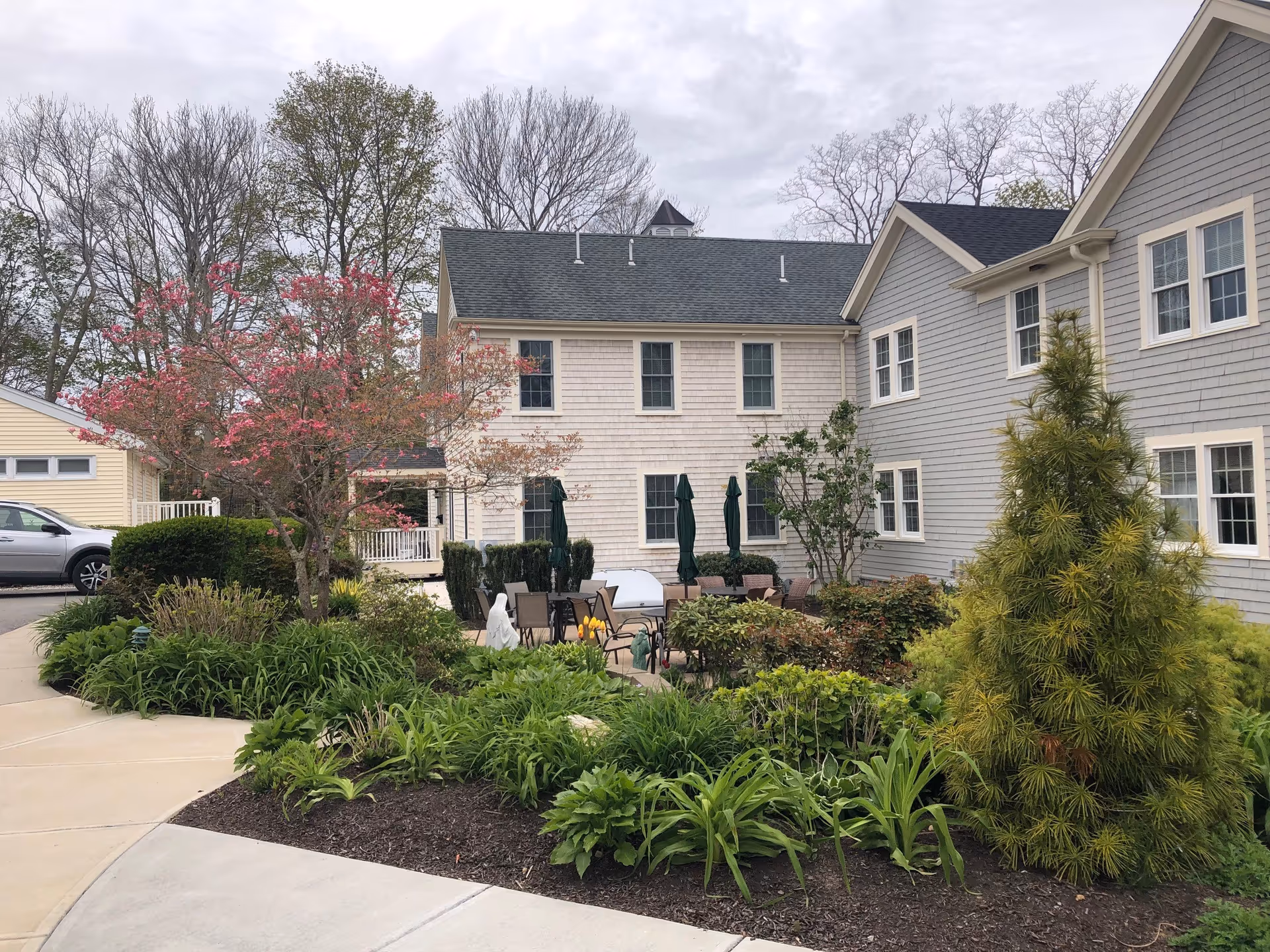 Outdoor garden area at Decatur House featuring a variety of green plants, bushes, and a tree with pink blossoms. There is a paved walkway curving around the garden, and patio furniture with closed umbrellas is visible near the building. The building exterior is light gray and beige with multiple windows, under a cloudy sky.