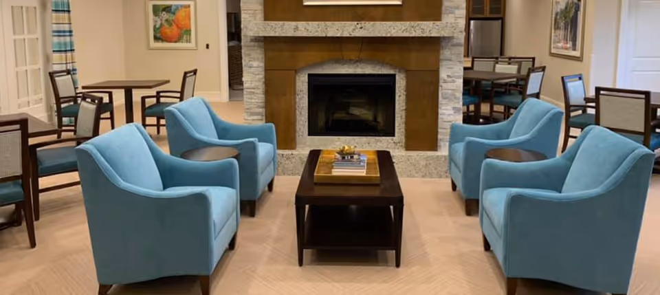 A cozy common area with four light blue armchairs arranged around a dark wooden coffee table with books on it, facing a stone fireplace with a wooden mantle. In the background, there are several dining tables with chairs, a painting on the wall, and a window with striped curtains.