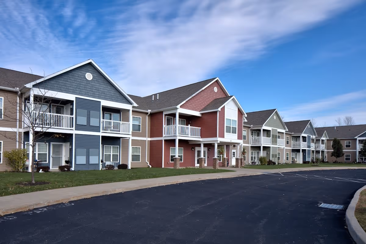 Exterior view of a senior living community building with multiple two-story units featuring balconies and porches. The building is painted in various colors including blue, red, beige, and green. There is a paved driveway and a small grassy area with a few trees in front of the building under a partly cloudy sky.