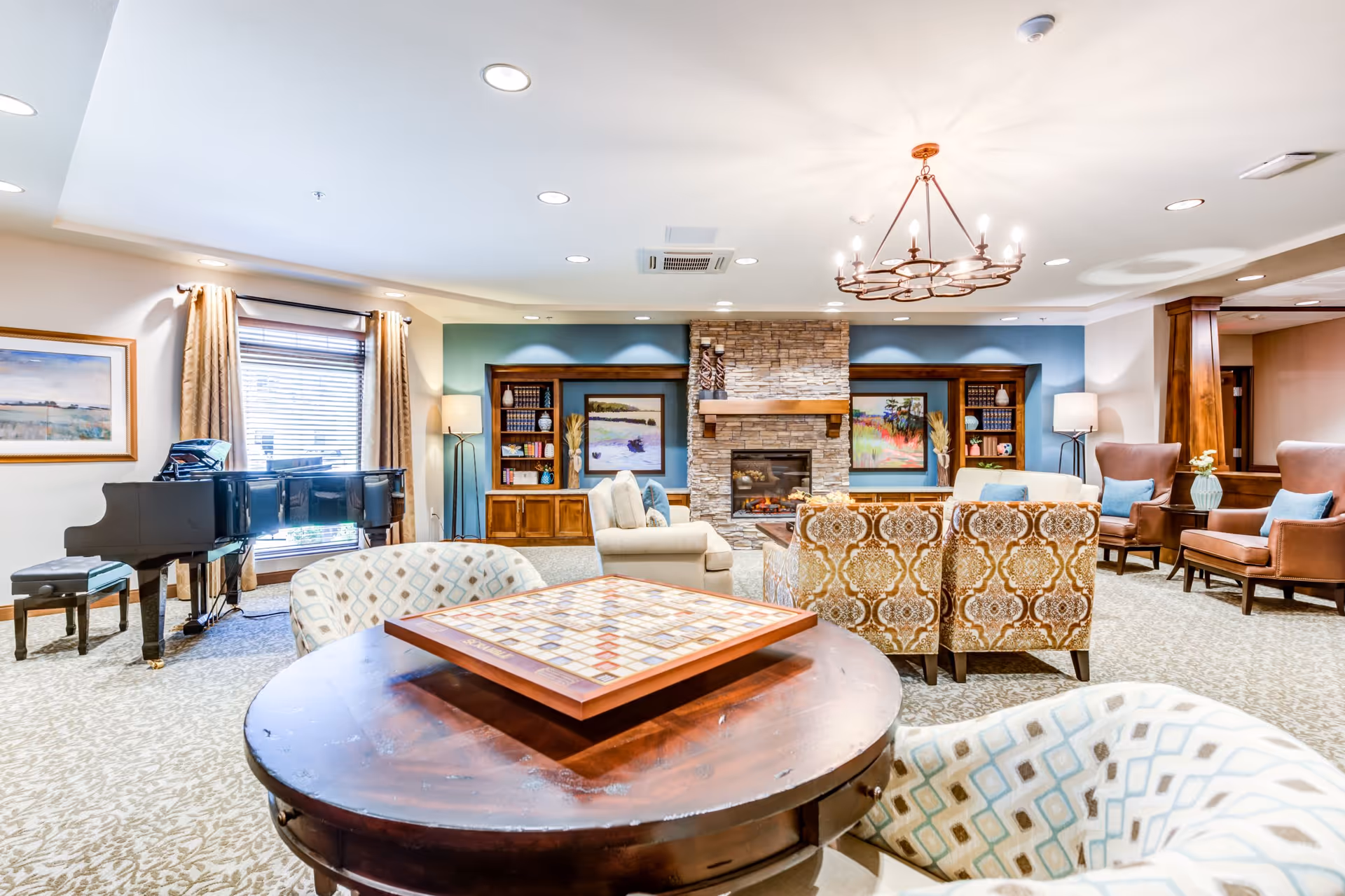 Spacious senior living common room with chairs, a stone fireplace, a grand piano, and a game table in the foreground.