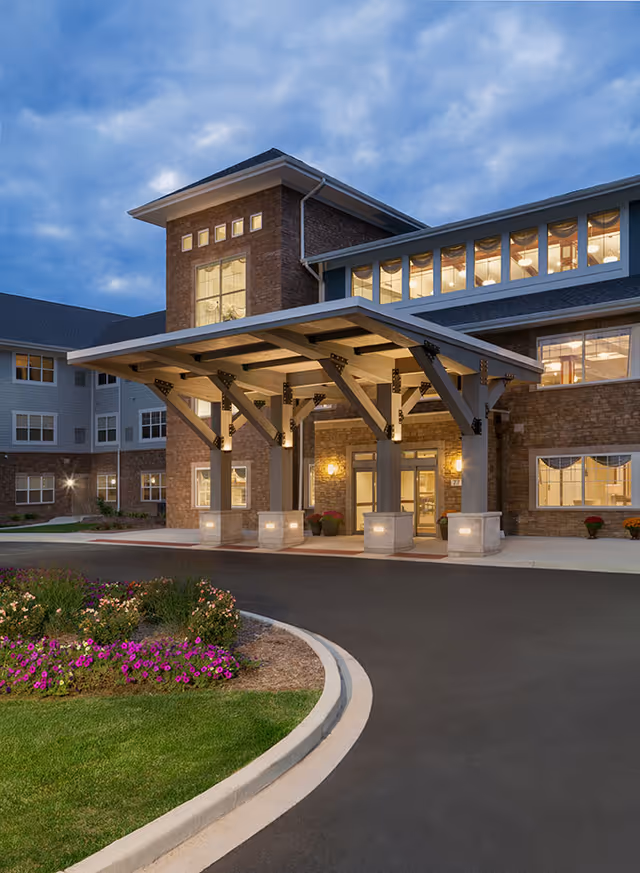 Exterior view of a senior living facility entrance at dusk with a covered drop-off area supported by large beams, illuminated lights, and a landscaped flower bed in the foreground.