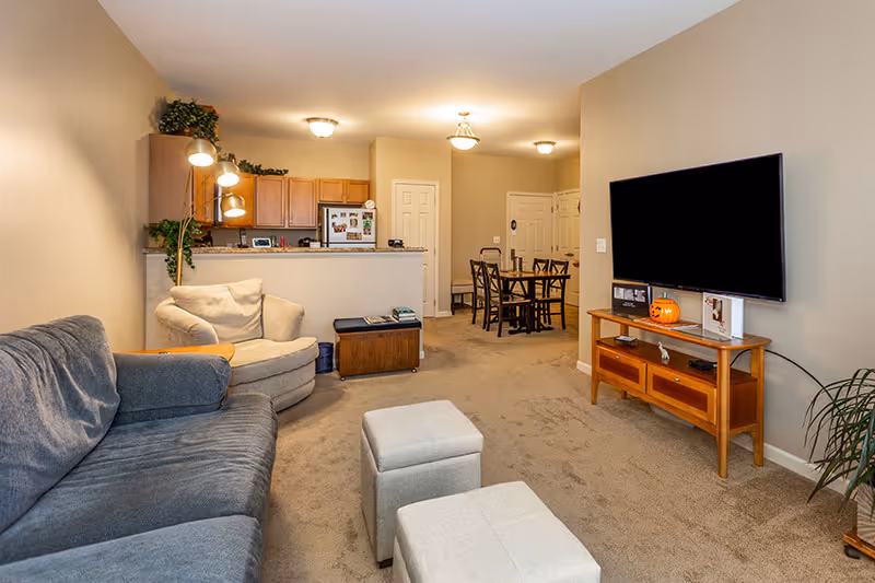 Interior view of a living room and dining area in an active adult retirement community. The living room features a gray sofa, a beige armchair, two small ottomans, a wooden TV stand with a flat-screen TV mounted on the wall, and some decorative plants. The dining area has a wooden dining table with chairs and is adjacent to a kitchen with wooden cabinets and a refrigerator.