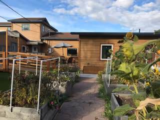 Outdoor garden area with raised garden beds filled with plants and flowers, a paved walkway leading to a wooden building under a partly cloudy sky.