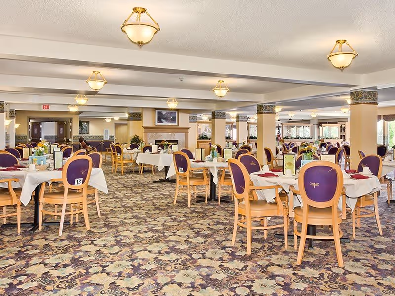 Large dining room with tables set for meals and purple-upholstered chairs in a retirement community.