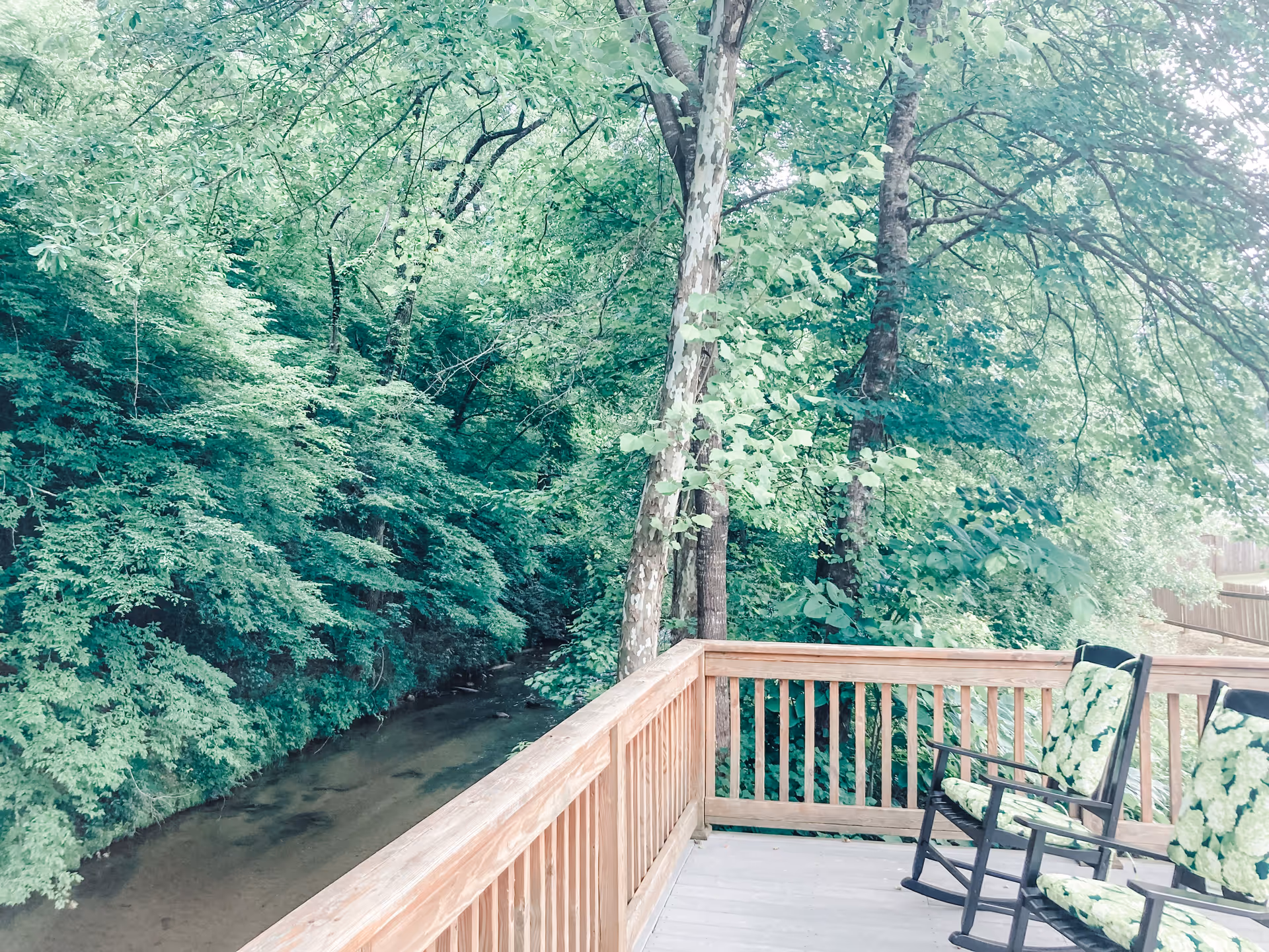 Wooden deck with two floral-cushioned rocking chairs overlooking a wooded creek and dense trees.