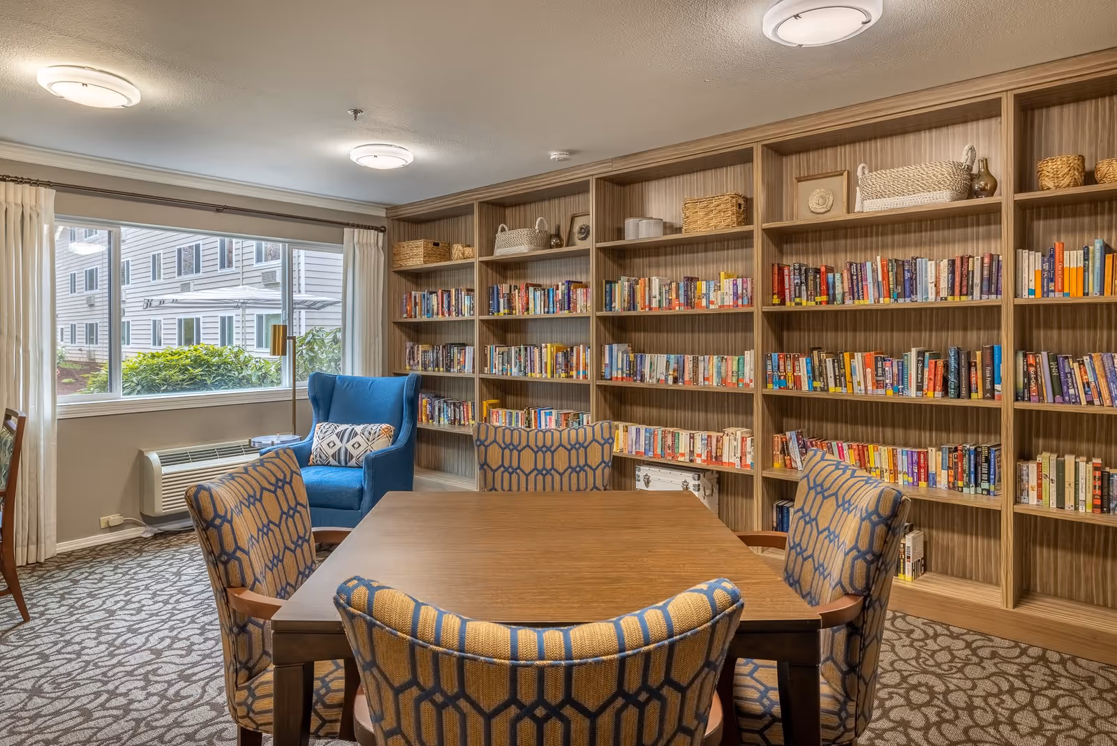 A cozy reading room in a retirement facility featuring a wooden table surrounded by four patterned chairs, a blue armchair with a decorative pillow near a large window with white curtains, and a wall lined with bookshelves filled with books and decorative baskets.