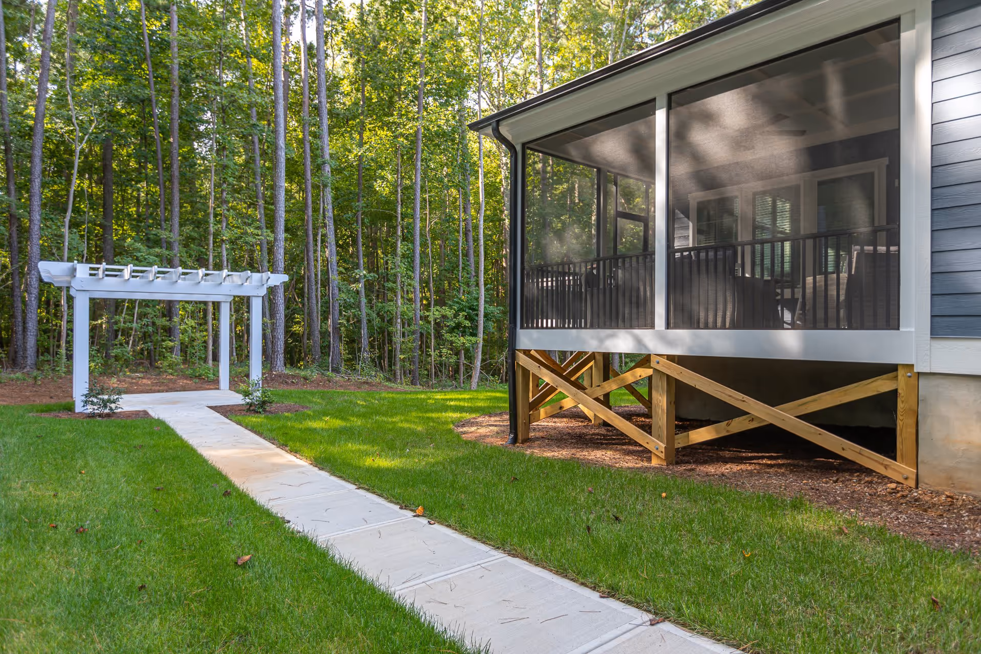 A paved walkway leads through a green lawn to a white wooden pergola with a backdrop of tall trees. To the right, there is a screened porch attached to a building with wooden support beams underneath.