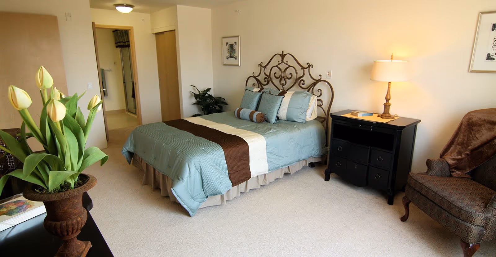 A neatly made bed with a decorative metal headboard in a bedroom. The bed has light blue bedding with a brown and white runner and several pillows. To the right of the bed is a black nightstand with a lamp and a book. There is a patterned armchair with a brown throw blanket on the right side of the room. A potted plant with yellow tulips is visible in the foreground on the left side. The room has beige carpet and light-colored walls with framed artwork.