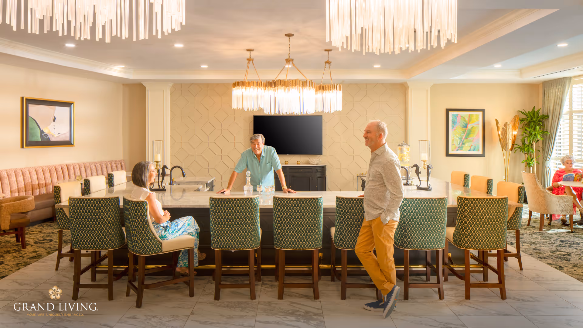 A bright and spacious common area in a senior living facility featuring a large marble island with green patterned chairs around it. Three seniors are interacting near the island, with one woman seated, one man standing, and another man leaning on the island. The room is decorated with modern chandeliers, framed artwork, plants, and comfortable seating areas with a carpeted floor and large windows allowing natural light.