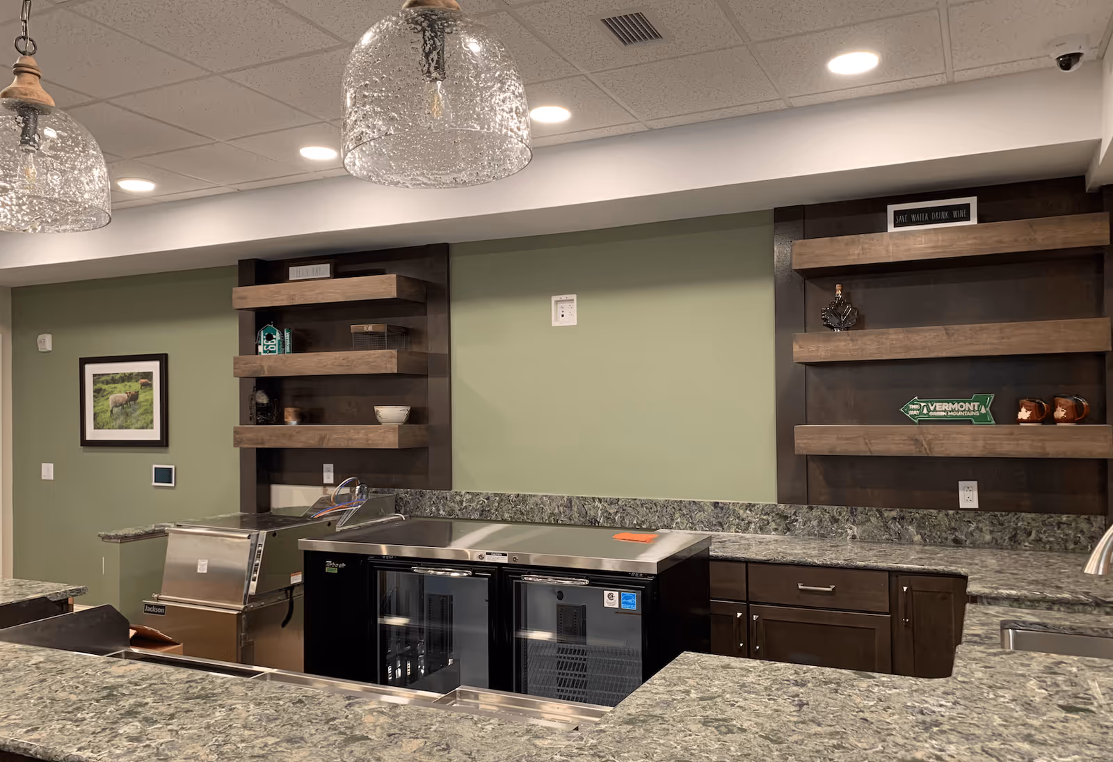 Interior view of a kitchen area with green granite countertops, dark wood cabinets, and open wooden shelves displaying decorative items. Two glass pendant lights hang from the ceiling, and there is a stainless steel sink and beverage coolers under the counter. The walls are painted light green.