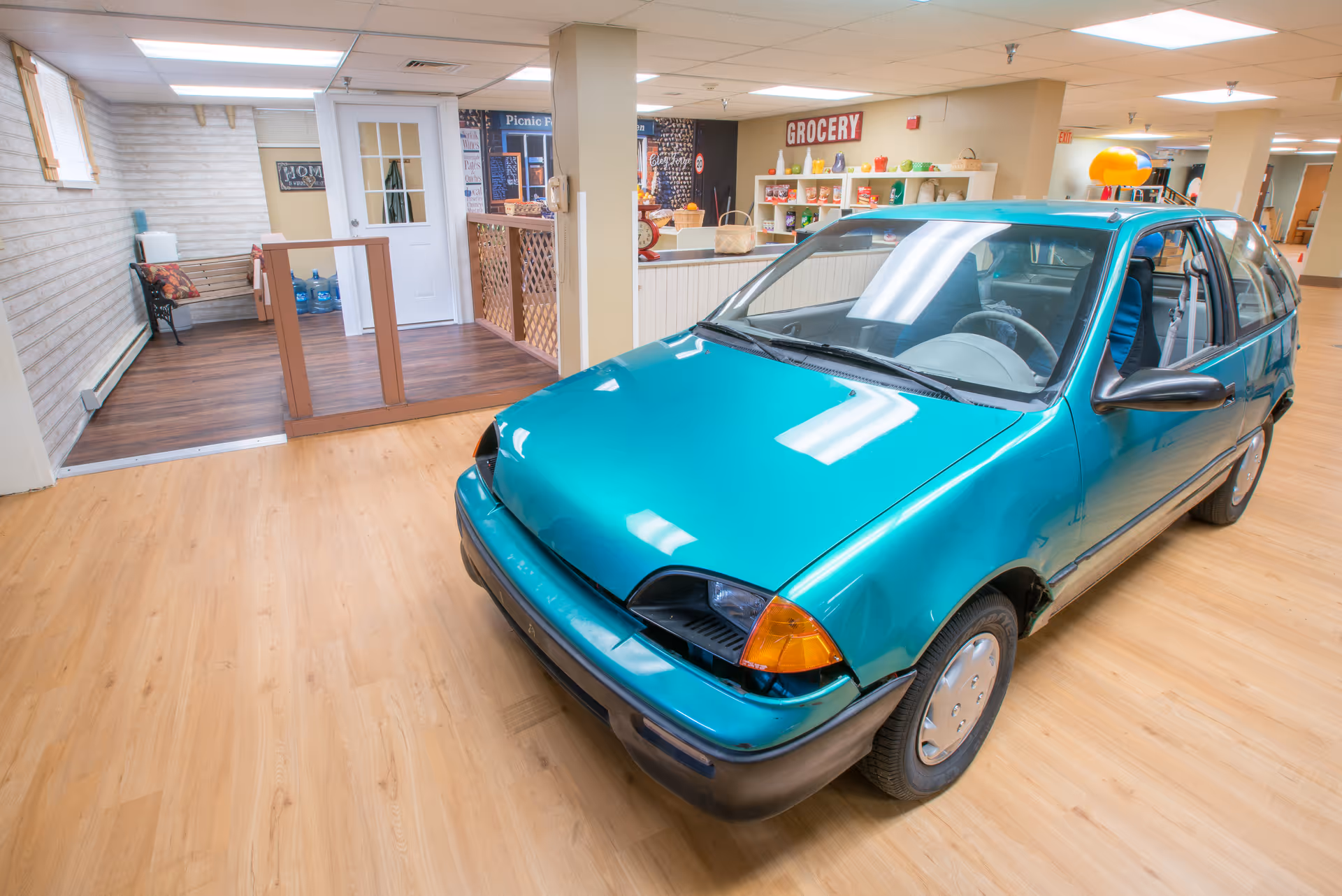 A teal compact car parked inside a nursing facility's indoor common area near a mock storefront labeled "GROCERY".