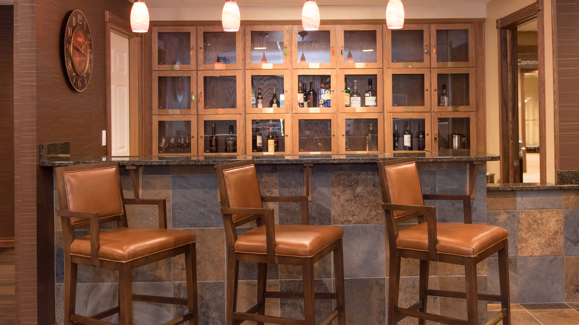 A cozy bar area with three brown leather bar stools in front of a granite countertop. Behind the bar is a wooden cabinet with glass doors displaying various bottles and glassware. Three pendant lights hang from the ceiling above the bar, and a large wall clock is mounted on the left wall.