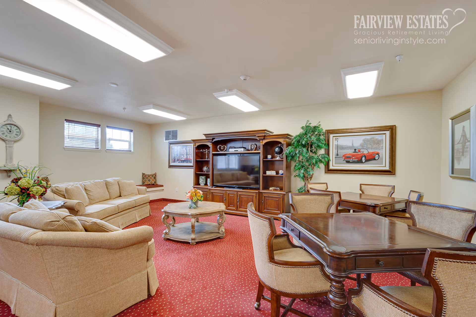 A cozy living room area with beige sofas, a wooden coffee table with a flower vase, a large wooden entertainment center with a TV, and a dining area with wooden tables and chairs. The room has red carpet flooring, beige walls, framed artwork, and ceiling lights. A potted plant is placed near the dining area.