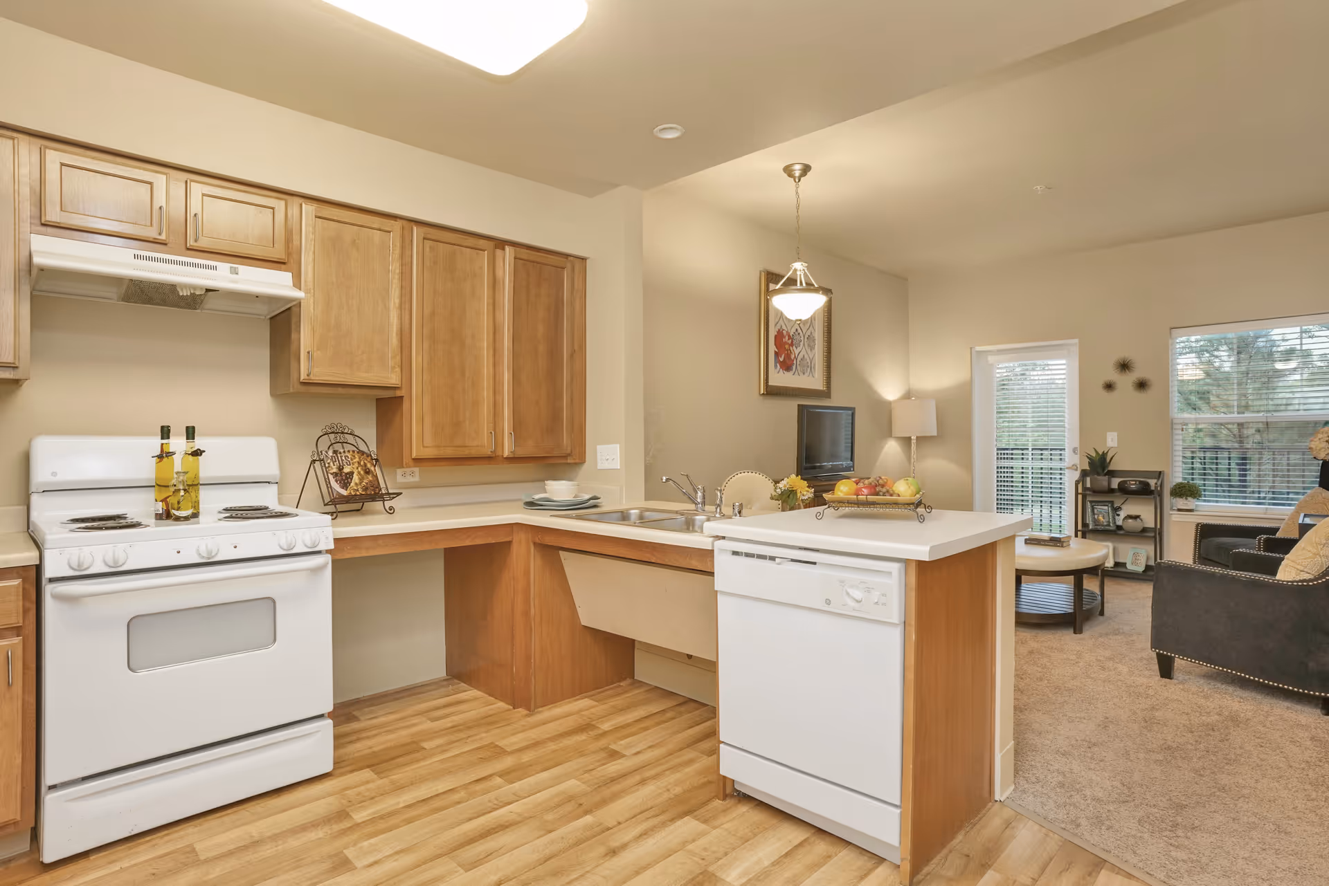 A bright kitchen area with wooden cabinets, a white stove, and a white dishwasher. The kitchen opens into a living room with carpeted flooring, a black armchair, a coffee table, a TV, and large windows with blinds letting in natural light.