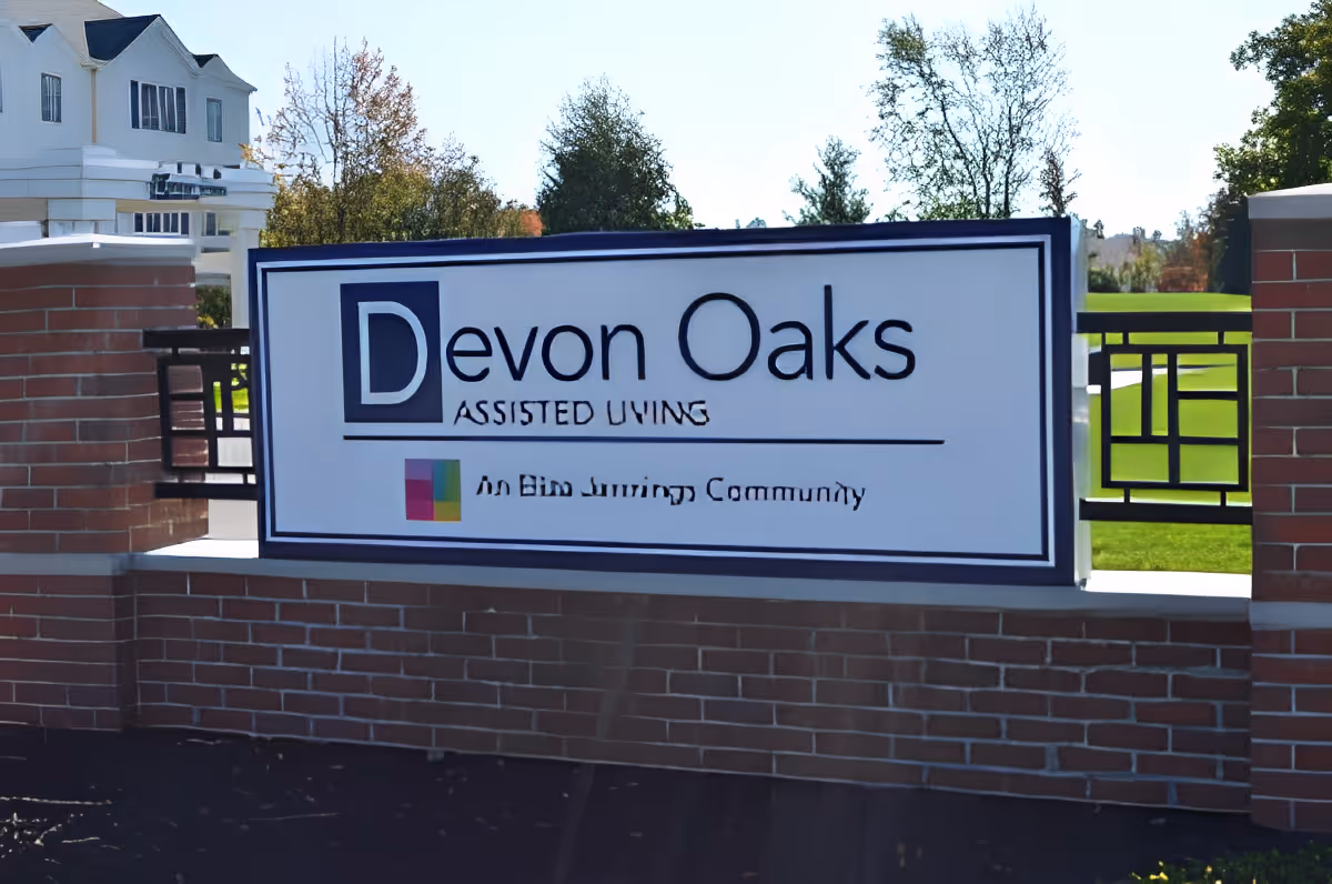 Entrance sign reading 'Devon Oaks Assisted Living' mounted on a brick wall with the facility and lawn visible behind it.