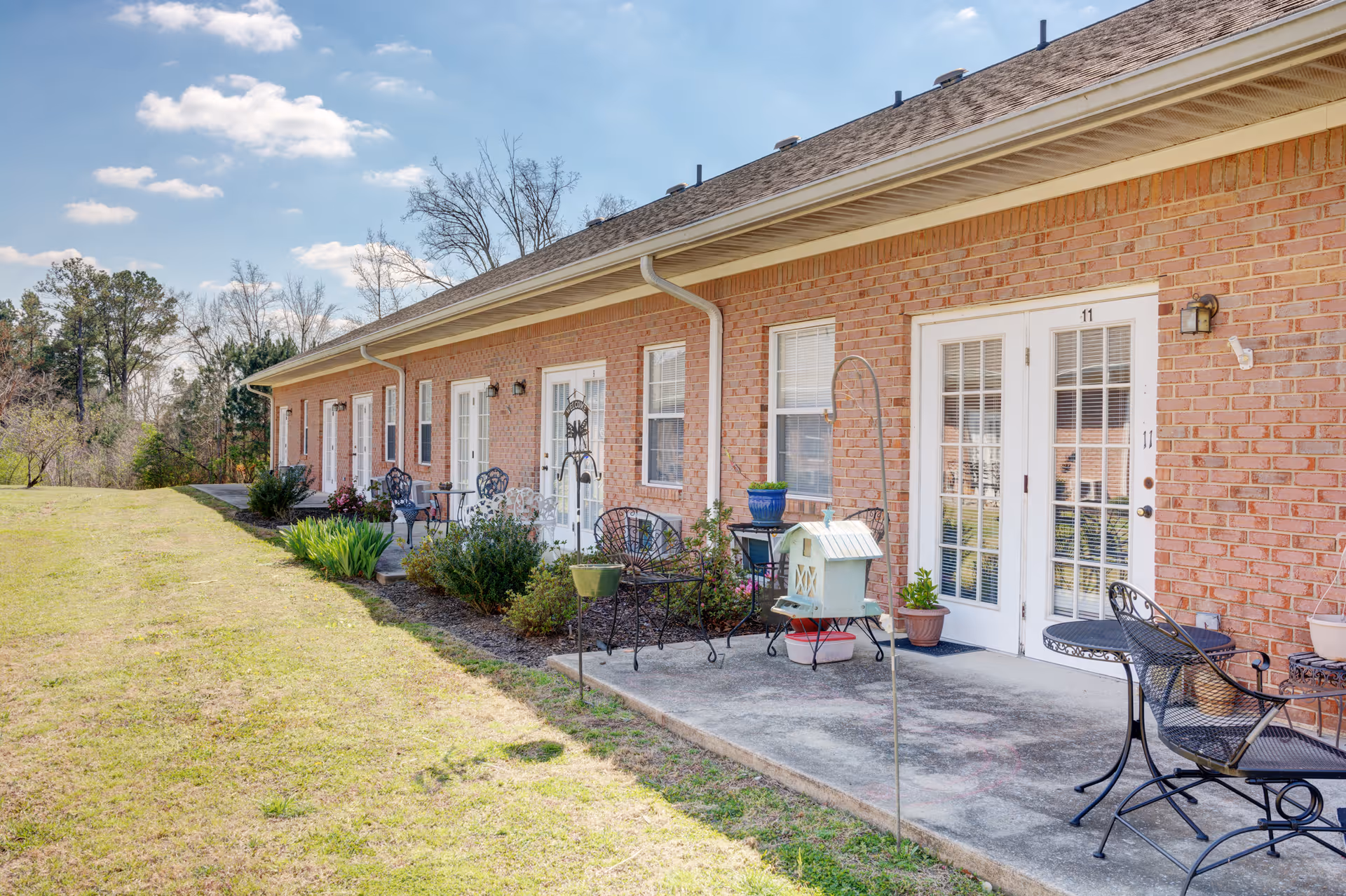Single-story brick senior living building with multiple patio doors, outdoor seating, and a grassy lawn.