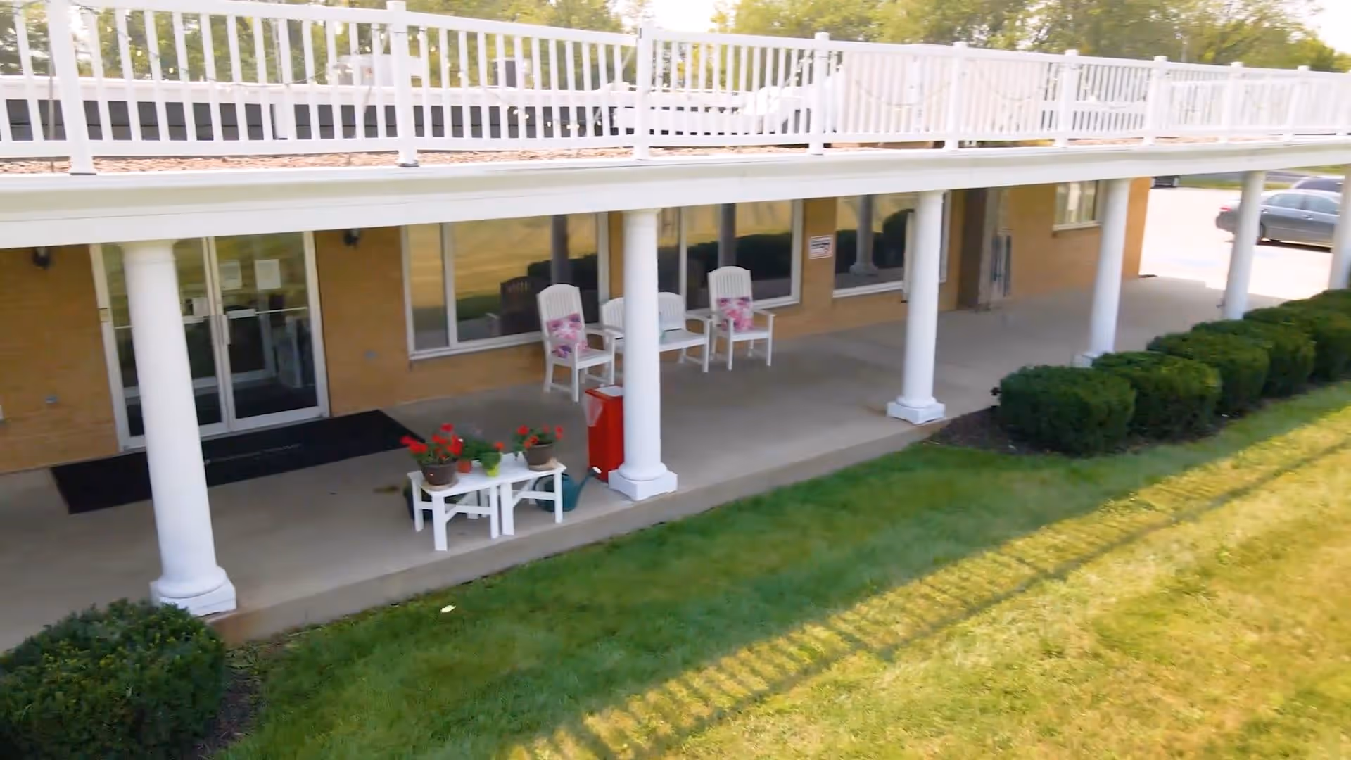 Outdoor covered patio area at Oakland Manor with white columns supporting an upper balcony. The patio has white chairs with floral cushions, a small white table with potted plants, and neatly trimmed bushes along the edge of the grass lawn.