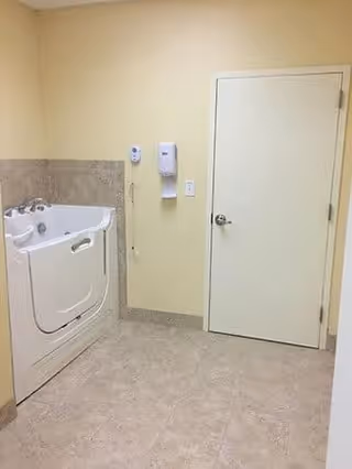 A clean, accessible bathroom featuring a walk-in bathtub with safety handles, beige tiled floor, light yellow walls, a closed white door, and mounted soap and sanitizer dispensers on the wall.