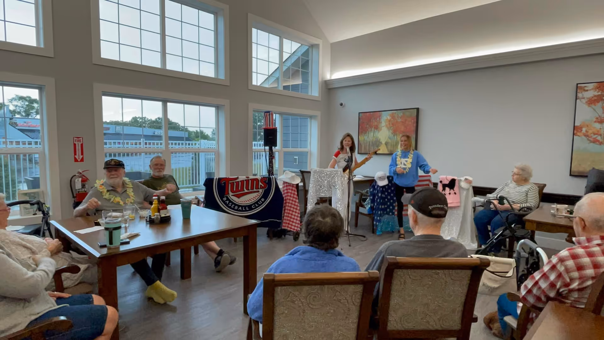 Residents seated around tables watch two performers singing and playing guitar in a bright common/activity room with tall windows.