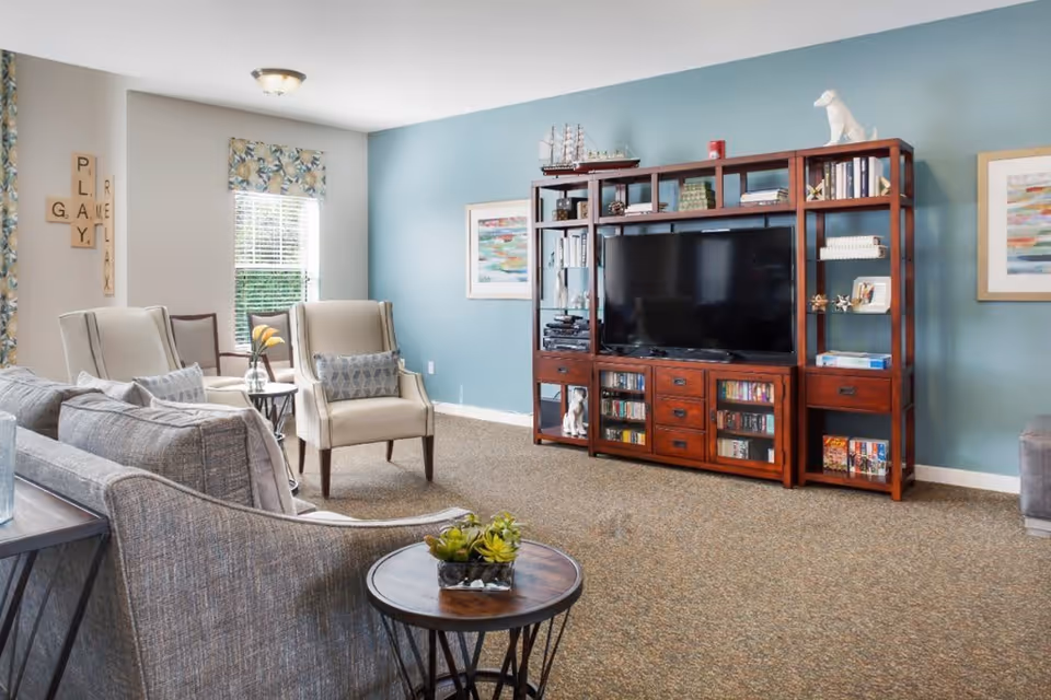 A cozy living room with a large wooden entertainment center holding a flat-screen TV, books, and decorative items. The room features a blue accent wall, two beige armchairs with patterned pillows, a gray sofa, a small round wooden side table with a plant, and windows with floral curtains letting in natural light.