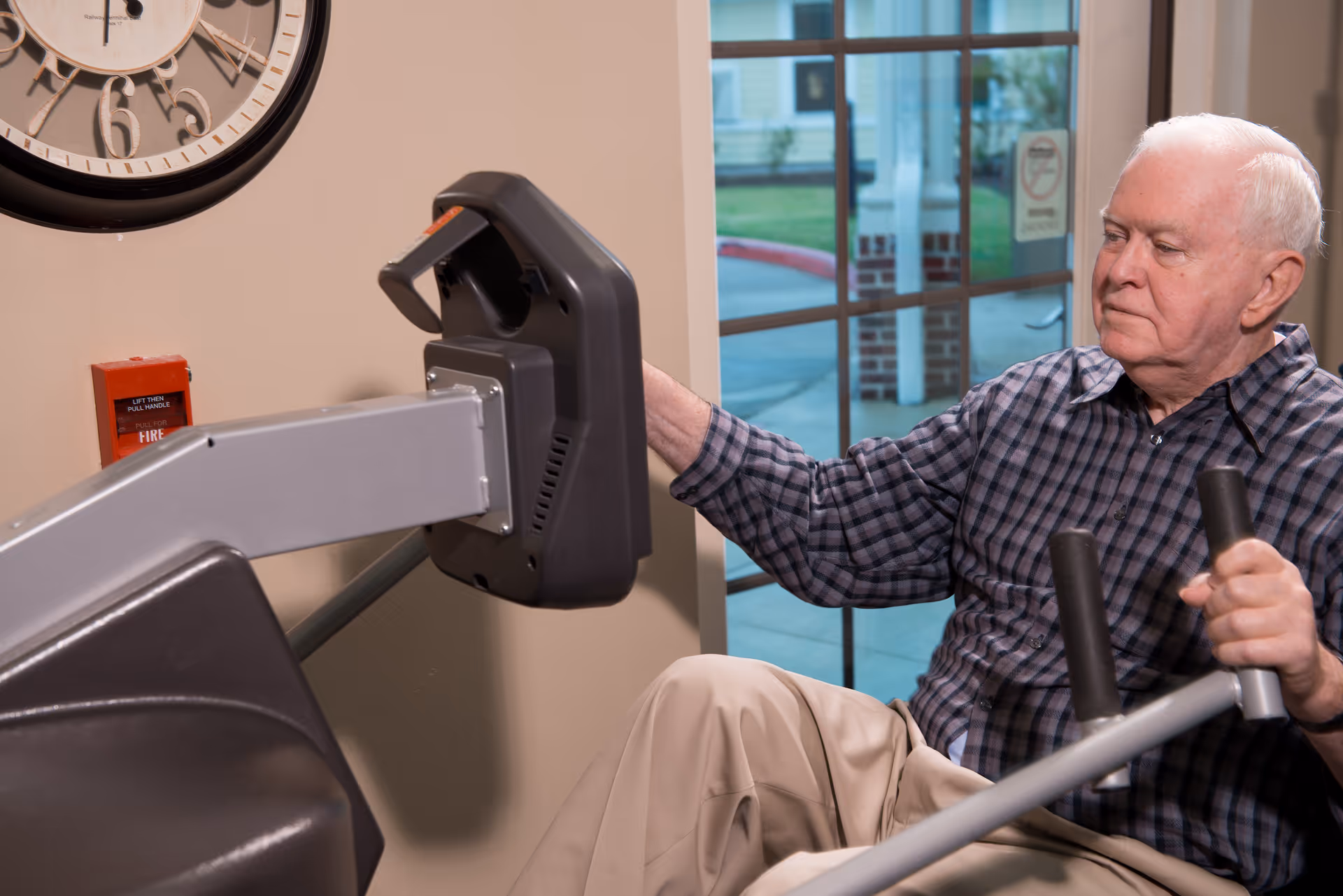 An elderly man wearing a checkered shirt and beige pants is using a seated exercise machine indoors near a large window. A wall clock and a fire alarm are visible on the wall behind him.
