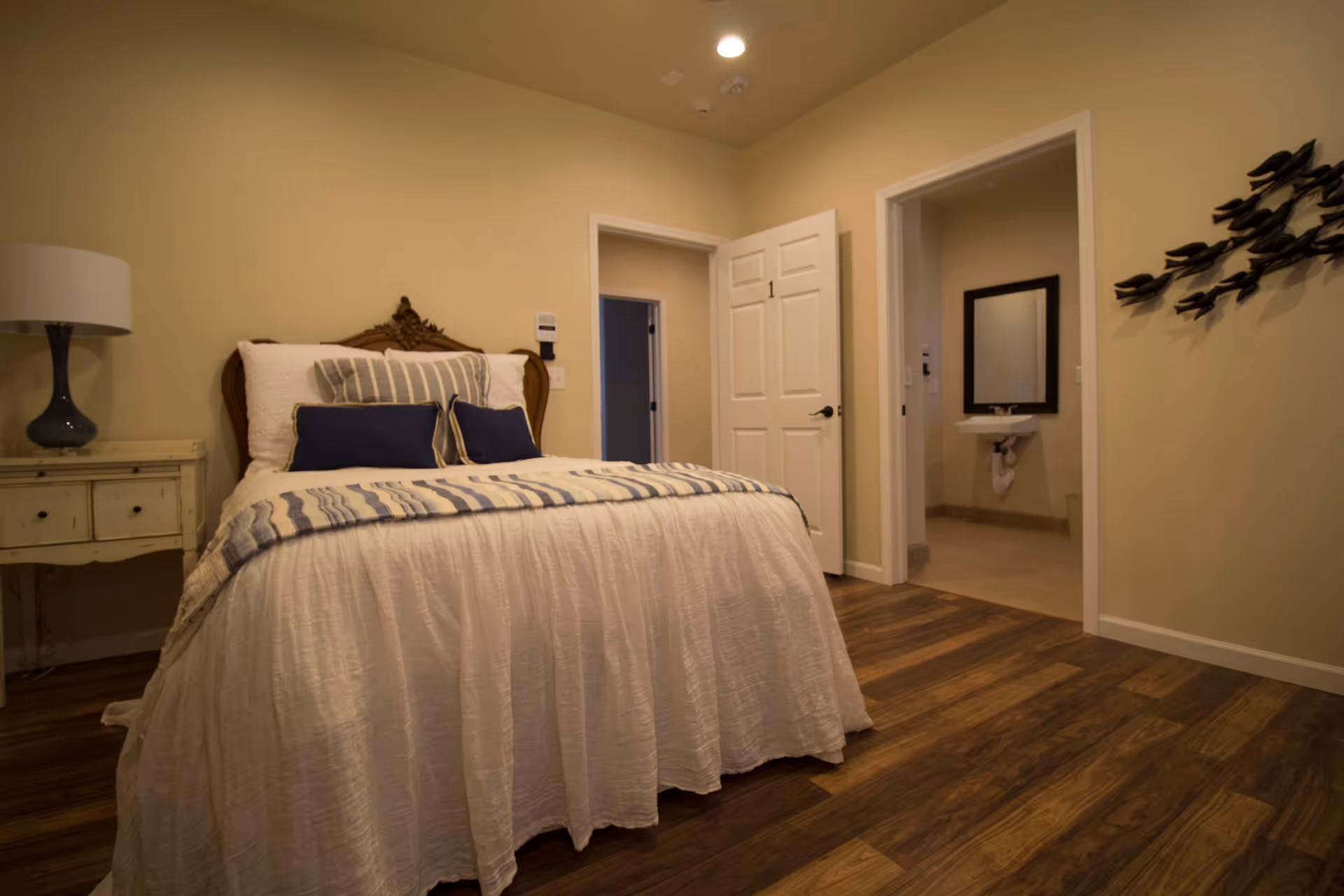 A cozy bedroom with a neatly made bed featuring white and blue bedding. There is a bedside table with a lamp on the left side. The room has wooden flooring and beige walls. Two open doors are visible, one leading to another room and the other to a bathroom with a sink and mirror.