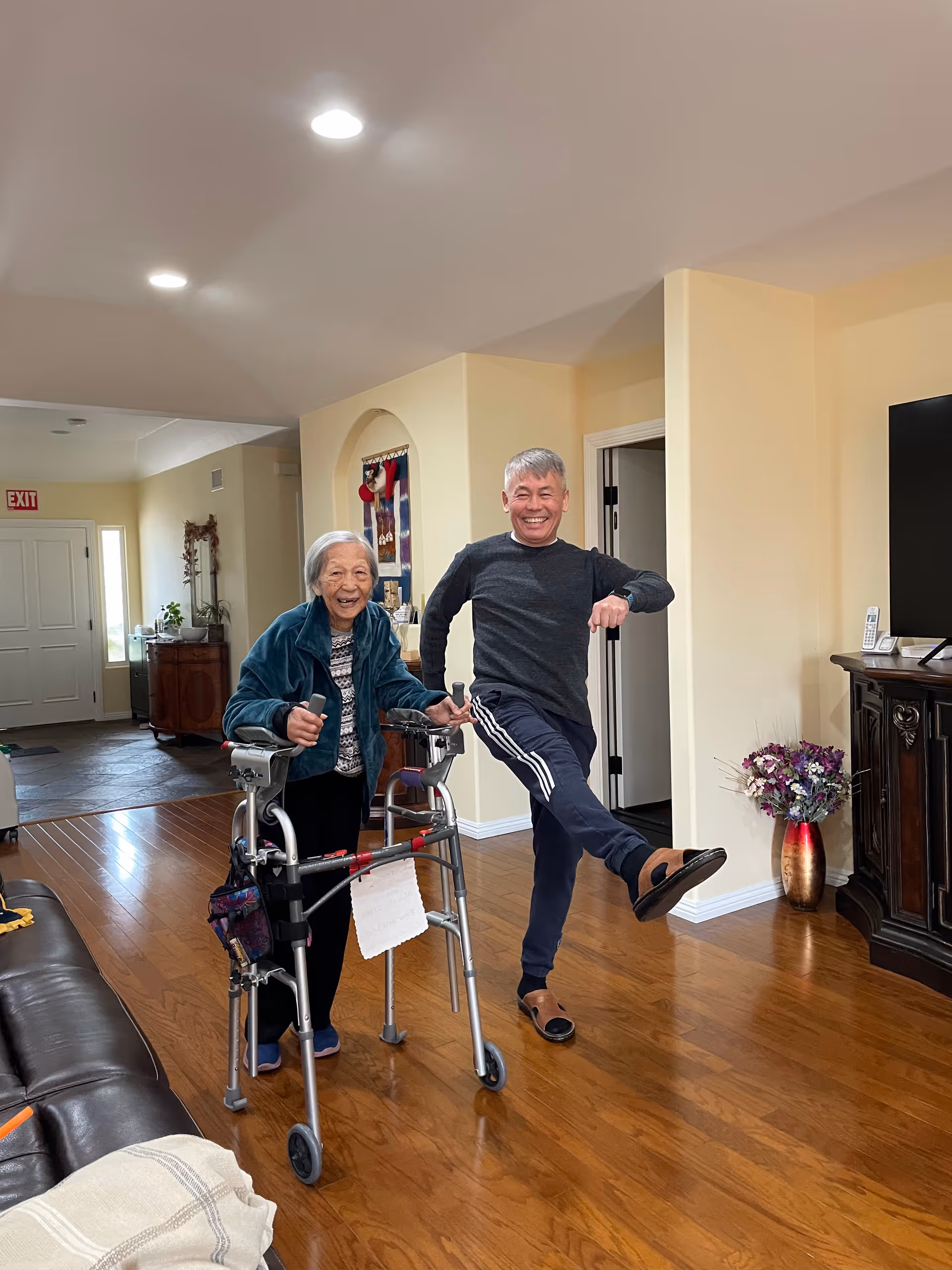 An elderly woman using a walker and a middle-aged man standing next to her, both smiling and posing playfully in a living room with wooden floors, a leather couch, a TV on a wooden stand, and a vase with flowers.