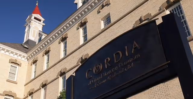 Exterior view of a large beige brick building with multiple windows and a white tower with a red roof. A large black sign on the building reads 'Cordia at Grand Traverse Commons'.