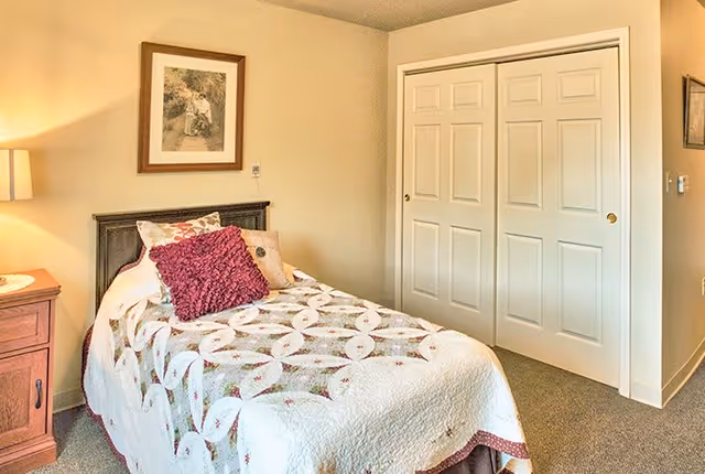 A cozy bedroom with a single bed covered in a patterned quilt and decorative pillows. There is a wooden nightstand with a lamp on the left side of the bed. A framed picture hangs on the wall above the bed. On the right side, there is a white double-door closet.