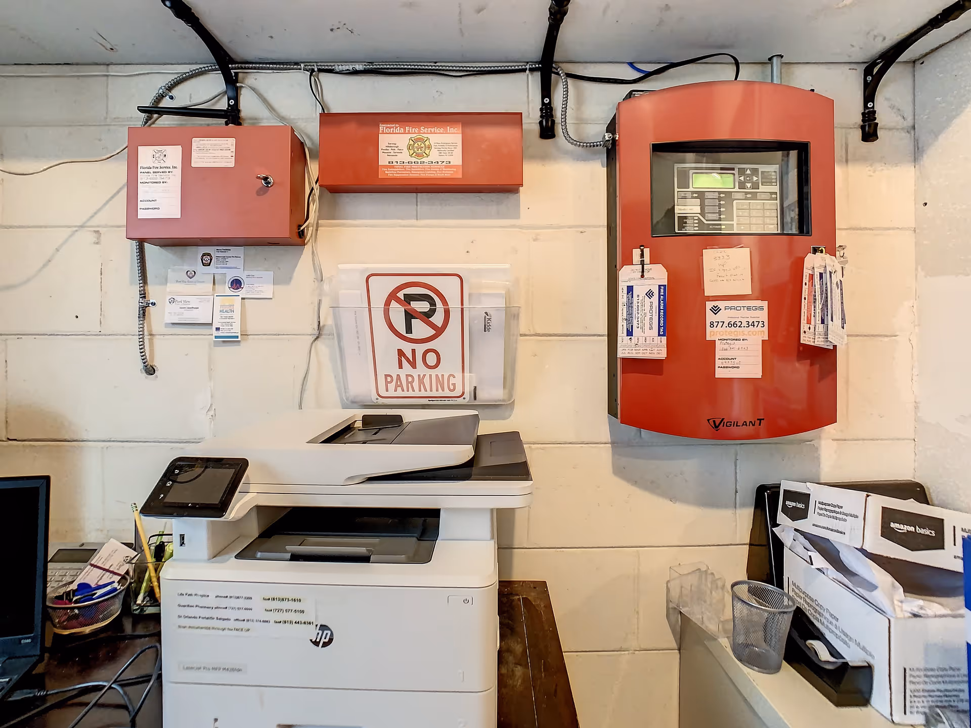 A utility room with a white HP laser printer on a wooden table, a computer monitor partially visible on the left, and various office supplies. On the wall above the printer are two red fire service control panels, a 'No Parking' sign in a clear holder, and several business cards and notes attached to the wall and panels. There is also a box labeled Amazon Basics on the right side of the image.