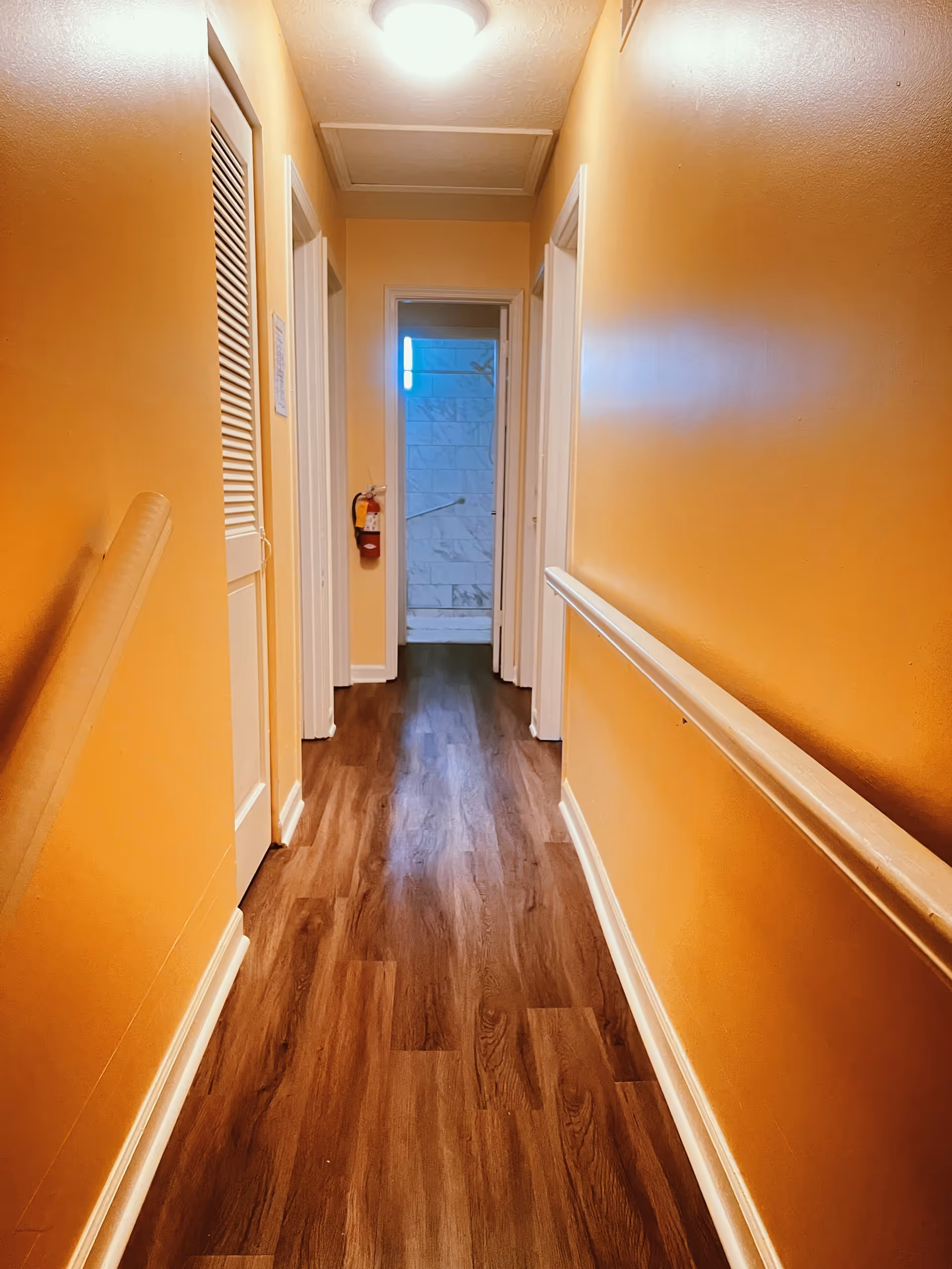 A narrow hallway with orange walls and wooden flooring leading to a bathroom with white tiled walls. There are white doors on both sides of the hallway and a fire extinguisher mounted on the left wall near the bathroom door.