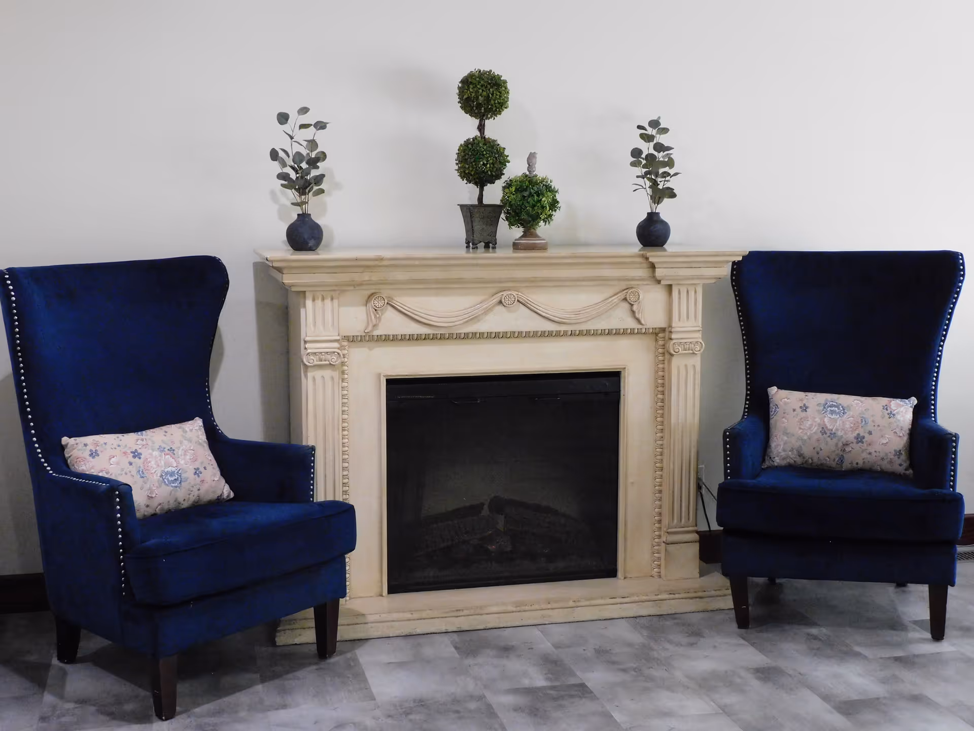 Two blue upholstered armchairs with floral cushions are positioned on either side of a decorative beige fireplace mantel. On top of the mantel are three small potted plants, including two with leafy branches and one with a topiary design. The floor is tiled in a gray pattern, and the wall behind is plain white.