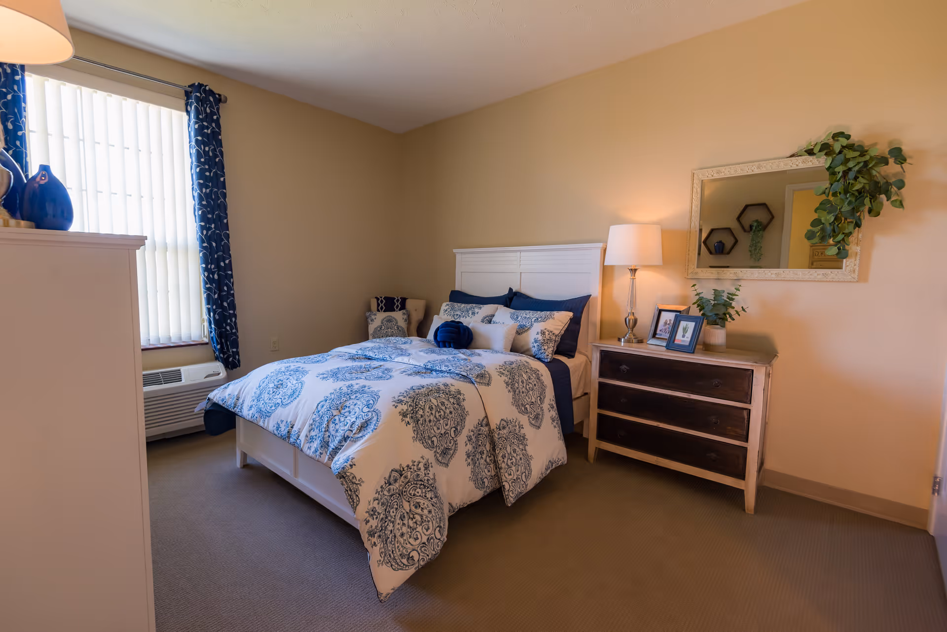A cozy bedroom with a white bed frame and a blue and white patterned comforter. There are multiple pillows on the bed, including navy blue and white ones. To the right of the bed is a wooden dresser with three drawers, a table lamp, framed pictures, and a potted plant. Above the dresser is a decorative mirror with a plant hanging on one corner. A window with vertical blinds and blue patterned curtains is on the left side of the room. The walls are painted beige and the floor is carpeted.