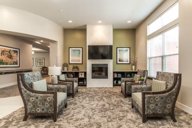 A cozy senior living community lounge area with four patterned armchairs arranged around a central fireplace and a wall-mounted flat-screen TV. The room features a large window with blinds, neutral-colored walls with two framed pictures, and built-in shelving on either side of the fireplace holding decorative items.