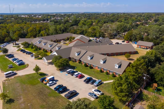 Aerial exterior view of a low-rise brick senior living complex with multiple wings, parking lots, lawns, and surrounding trees.