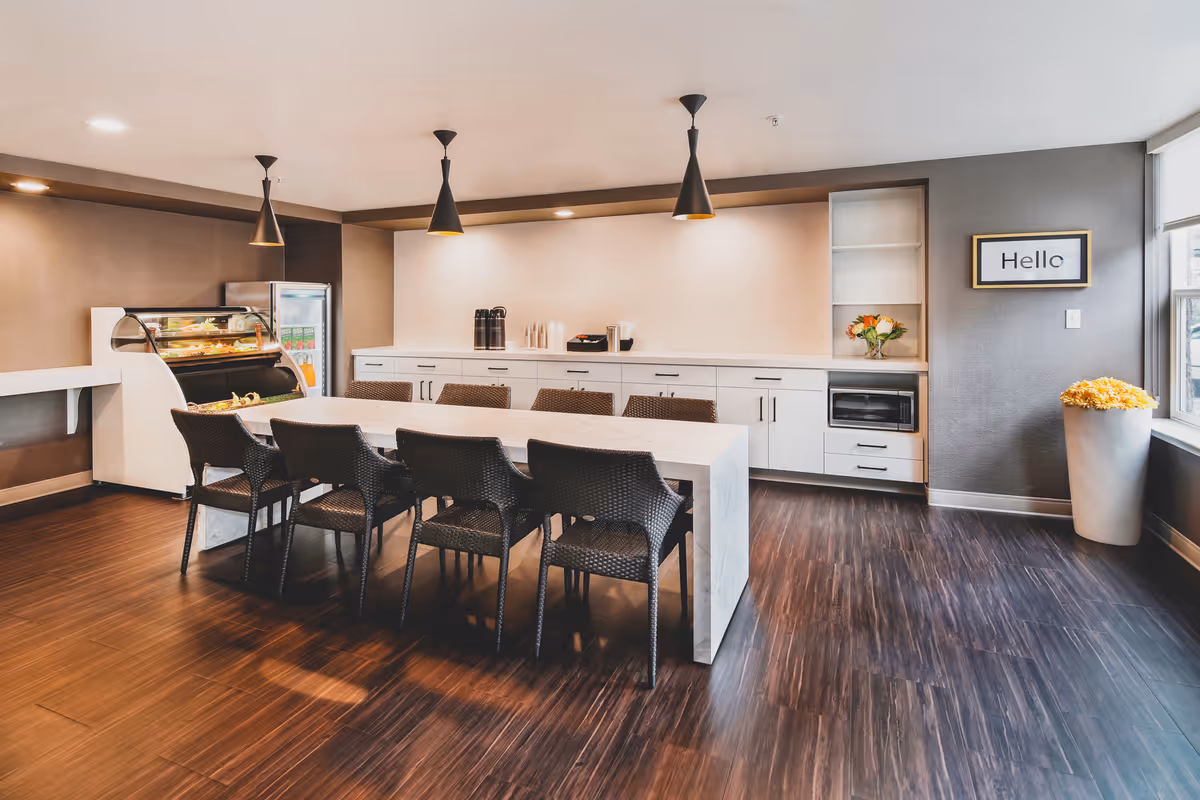 Bright communal dining area with a long marble table surrounded by wicker chairs, pendant lights, cabinets, and a refrigerated display case.