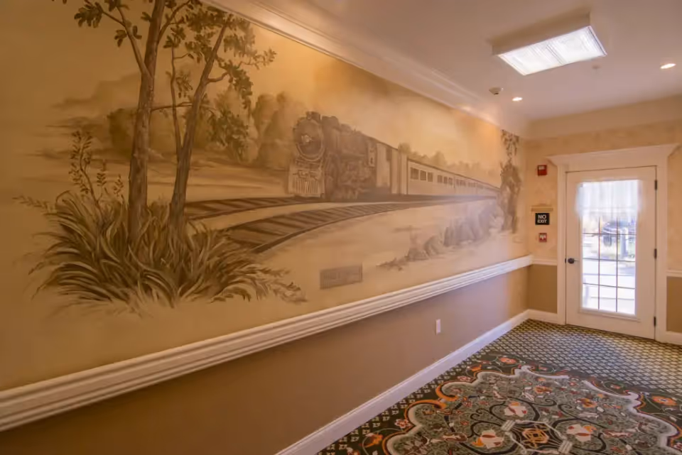 Interior hallway of an assisted living facility with a large mural of a vintage steam train on the wall. The hallway has patterned carpet, beige walls with white trim, a door with glass panes and a small curtain, and a 'No Exit' sign next to the door.